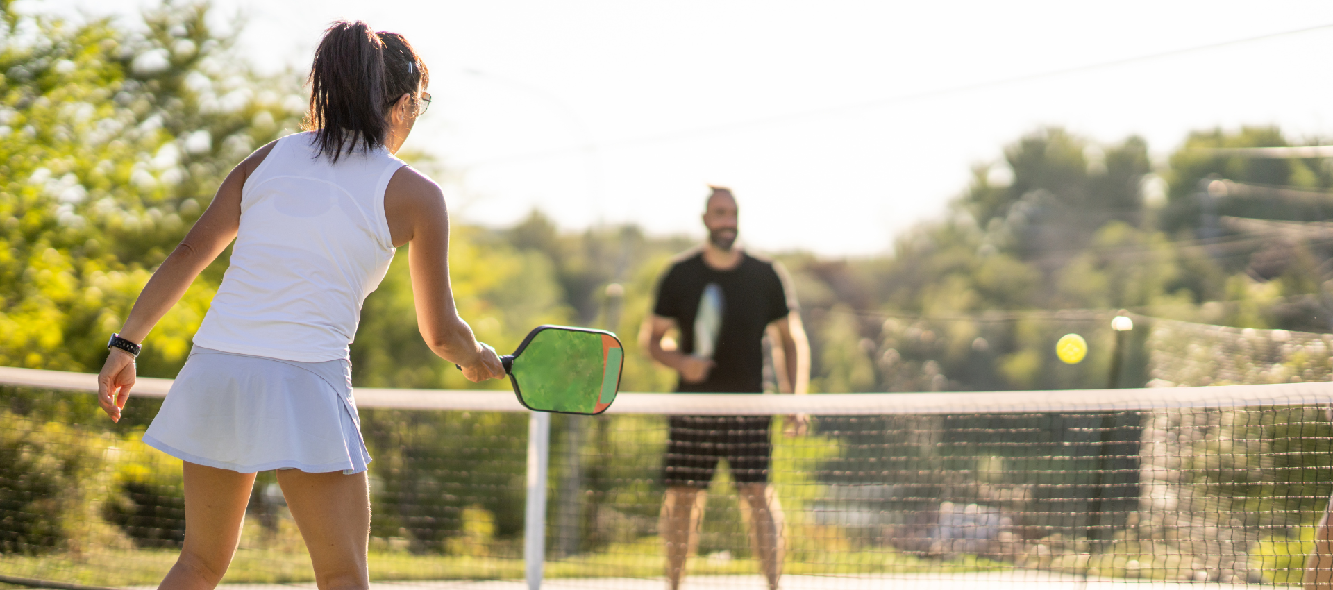 A woman and a man play pickleball outdoors on a sunny day, with the woman in the foreground ready to hit the ball over the net.