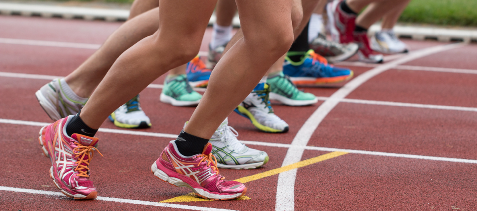 Runners wearing colorful athletic shoes line up at the starting line on a track, preparing for a race.