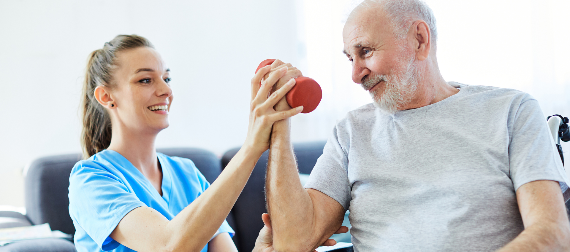 A nurse assists an elderly man in lifting a red dumbbell during a physical therapy session.