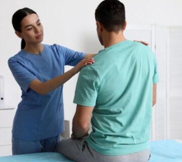 A healthcare professional in scrubs examines a seated male patient by gently touching his shoulders, demonstrating attentive care typical of Physical Therapy in Medford 