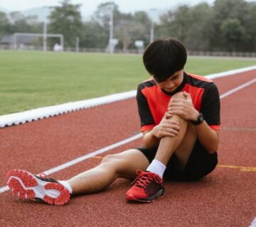 A young man in athletic clothing sits on a running track, holding his knee and appearing to be in pain—he may be looking for Physical Therapy in Medford to recover