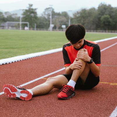 A young man in athletic clothing sits on a running track, holding his knee and appearing to be in pain—he may be looking for Physical Therapy in Medford to recover 