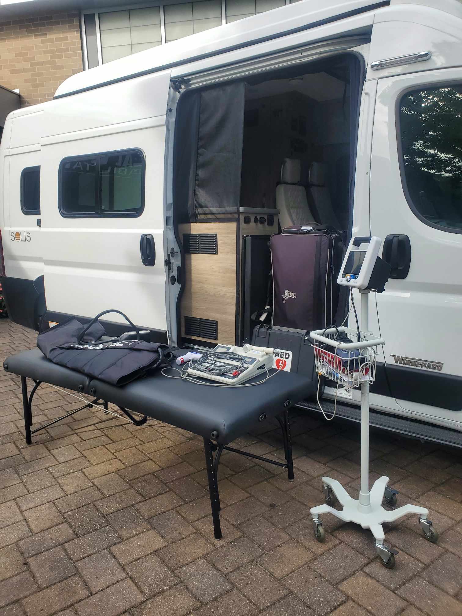 A medical van with its side door open is parked on a brick driveway  In front, there is a portable exam table with medical equipment and a rolling vital signs monitor 