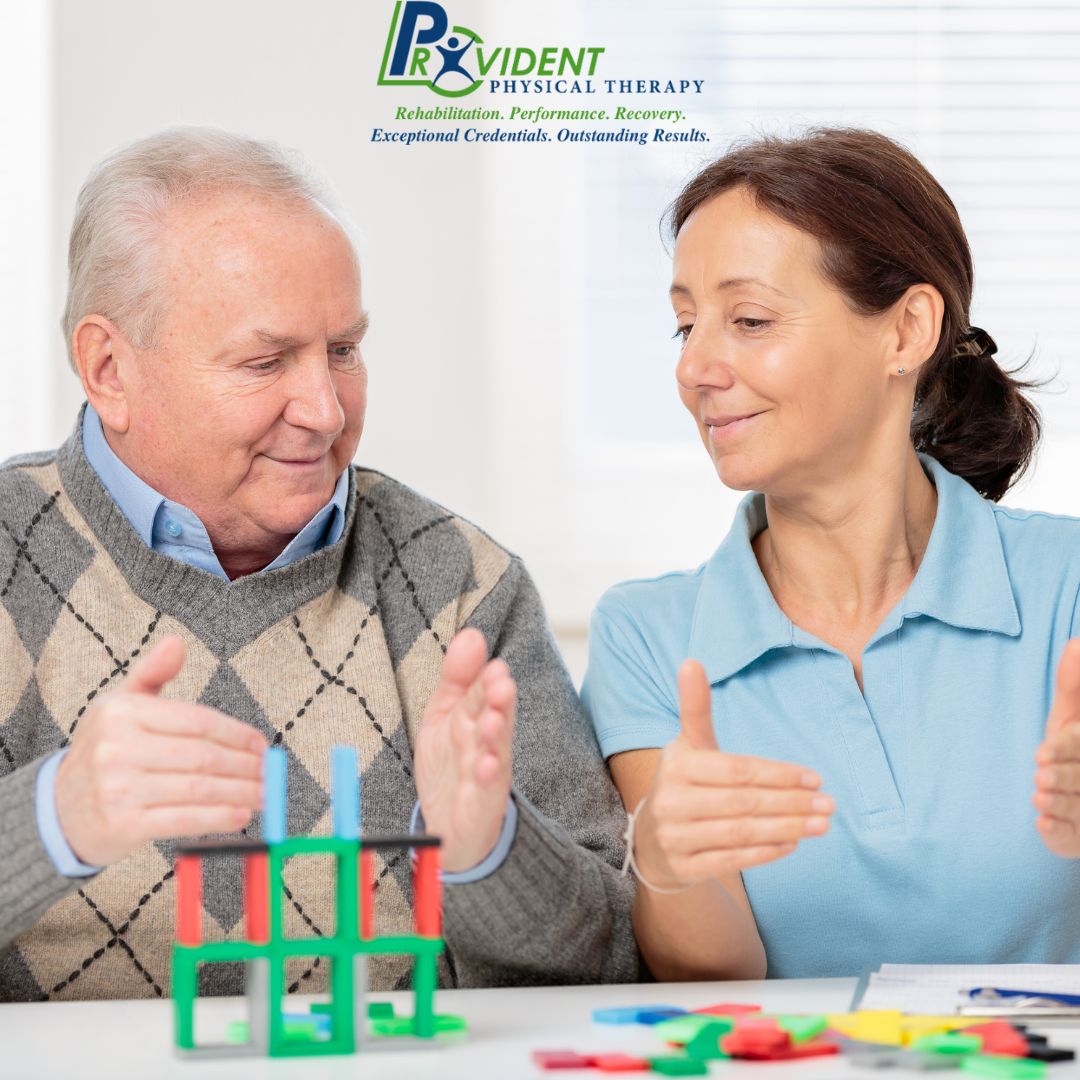 An older man and a female therapist work with colorful building blocks at a table during an occupational therapy session in Coram, NY  The Provident Physical Therapy logo is visible above them 