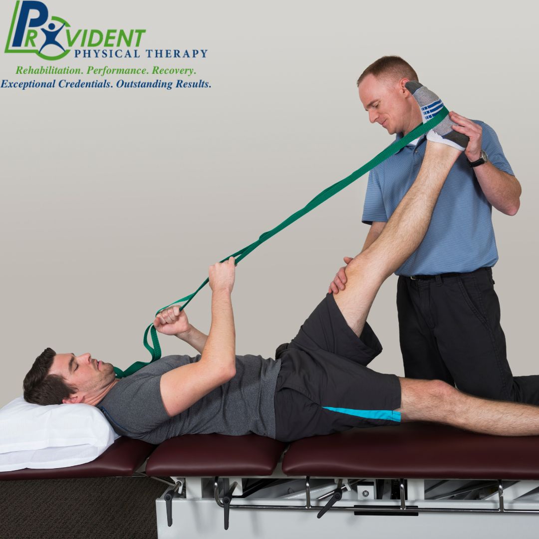 A physical therapist at Provident Physical Therapy in Medford stretches a patient’s leg using a green strap while the patient lies on a treatment table 