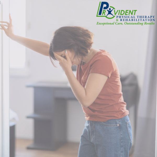 Woman standing indoors, leaning against a wall with one hand and covering her face with the other, appearing to experience discomfort from balance disorders; Provident Physical Therapy & Rehabilitation logo in the corner
