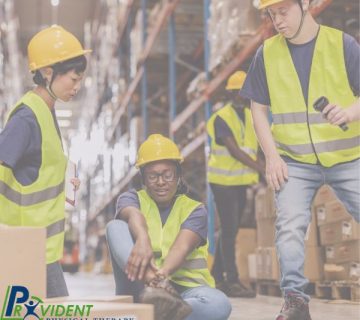 A warehouse worker sits on the floor holding her ankle while two colleagues in safety vests and helmets check on her  Provident Physical Therapy & Rehabilitation logo is visible 