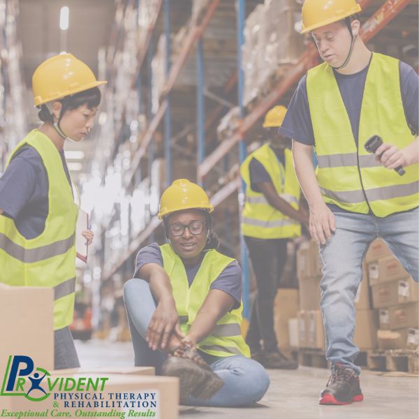A warehouse worker sits on the floor holding her ankle while two colleagues in safety vests and helmets check on her  Provident Physical Therapy & Rehabilitation logo is visible 